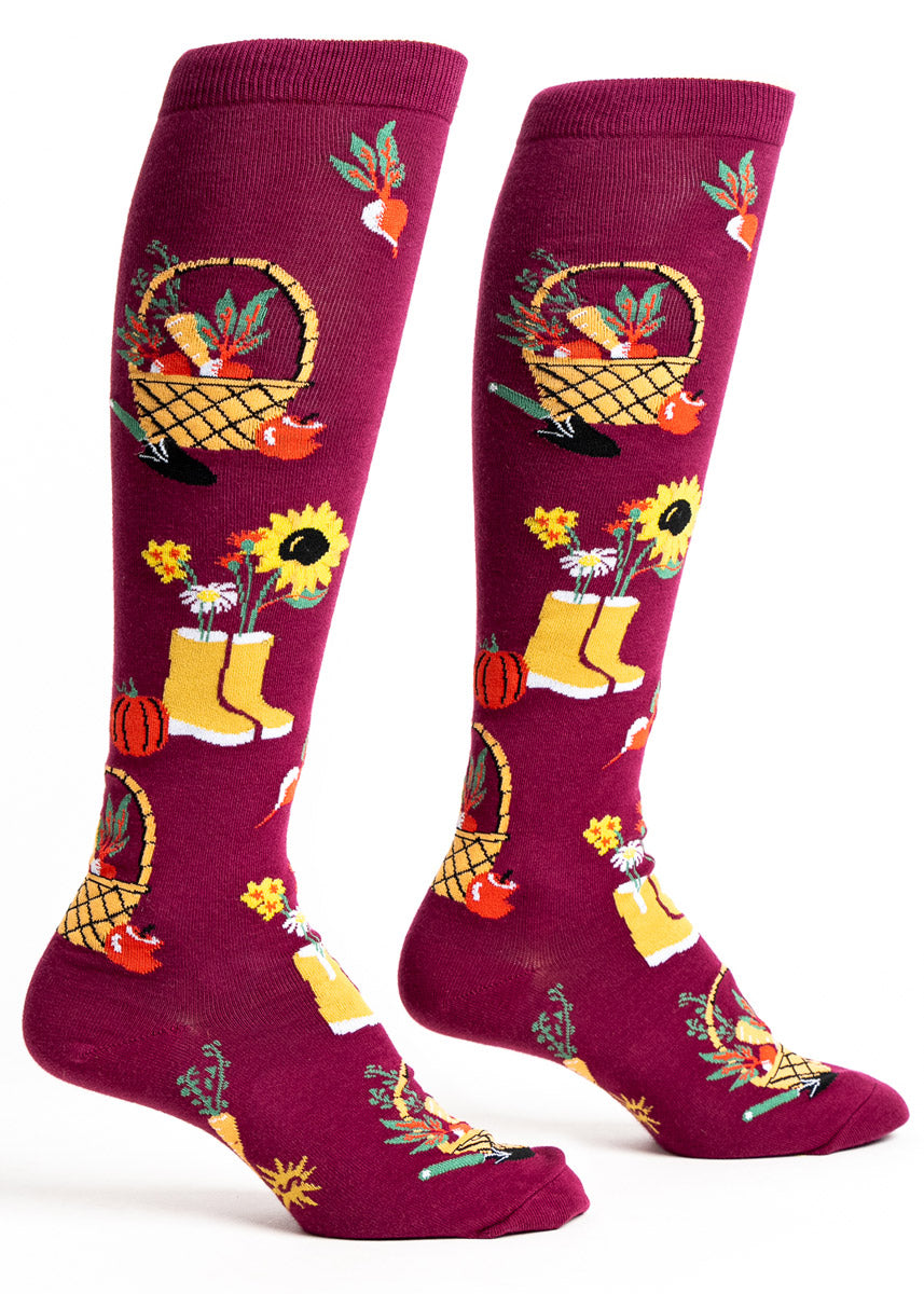 Maroon knee socks for women with a pattern of baskets of fall produce such as pumpkins and carrots, along with rain boots with sunflowers sticking out of the top.