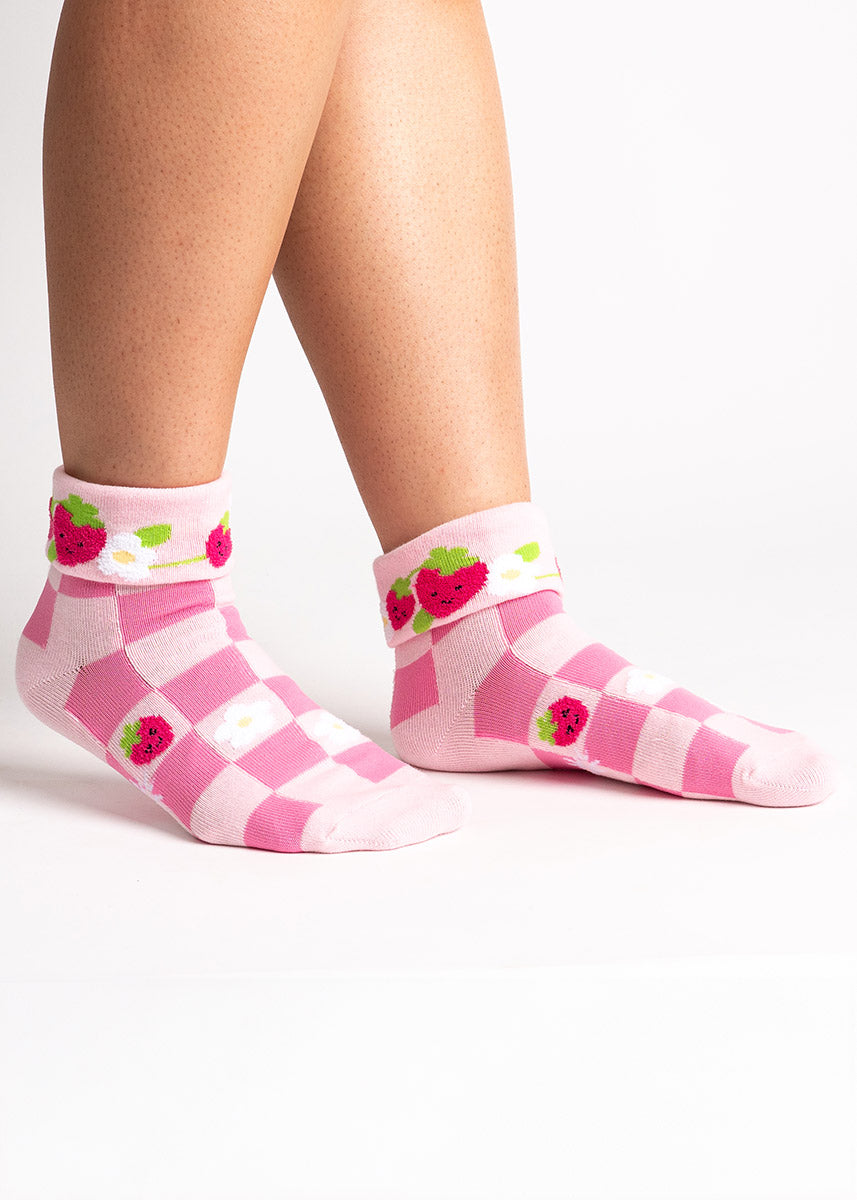 A female model poses wearing pink turn-cuff crew socks for women with an allover pattern of checkered print, with fuzzy strawberries and flowers.