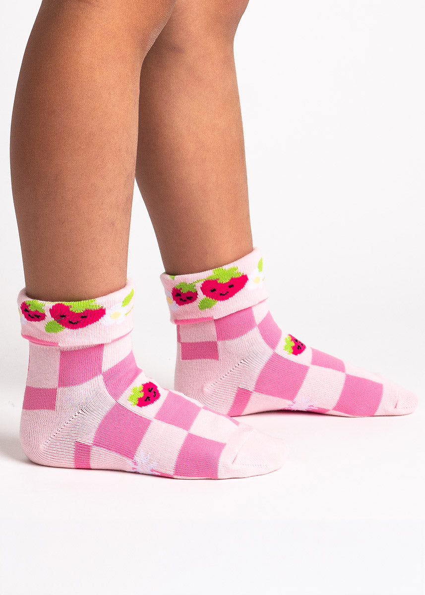 A child model poses wearing pink turn-cuff crew socks for kids with an allover pattern of checkered print, with fuzzy strawberries and flowers.
