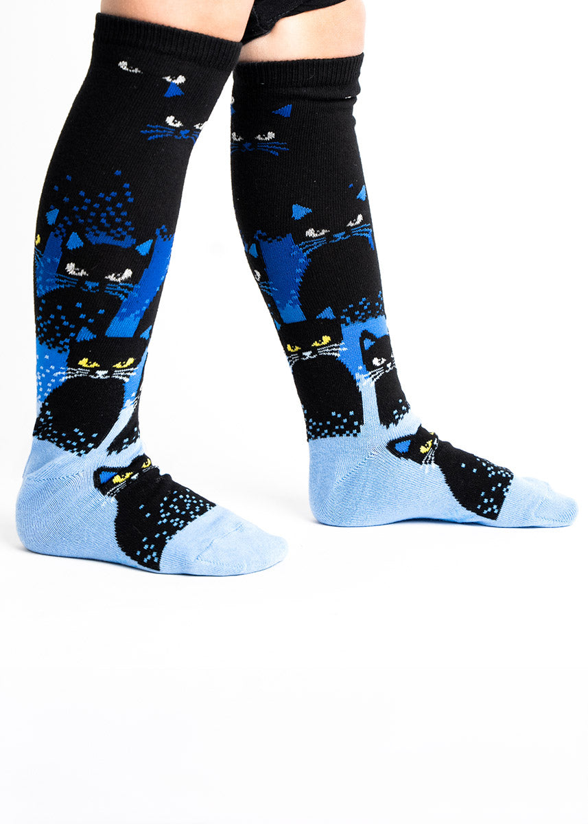 Knee socks for kids with a black-to-blue background gradient behind a pattern of black cats, the design creating the illusion of cats disappearing into the darkness.