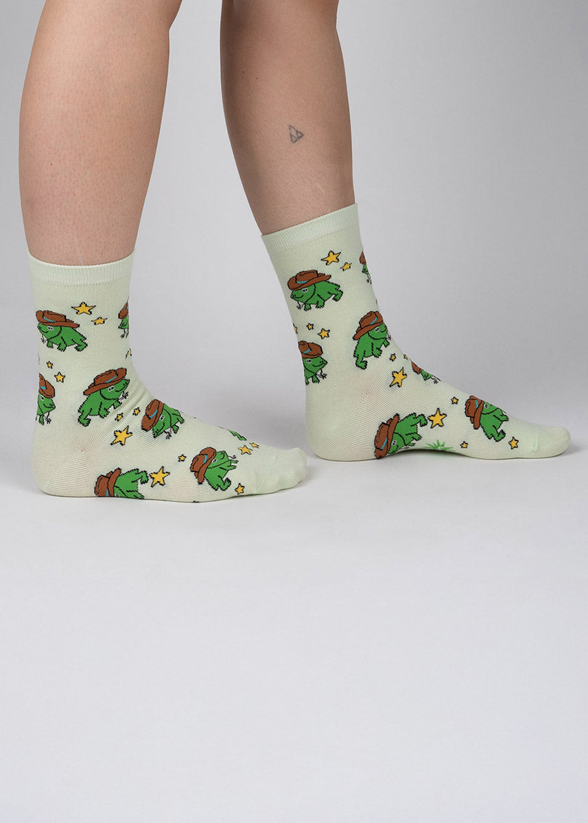 Light green crew socks for women with an allover pattern of green cartoon frogs wearing cowboy hats surrounded by yellow stars.