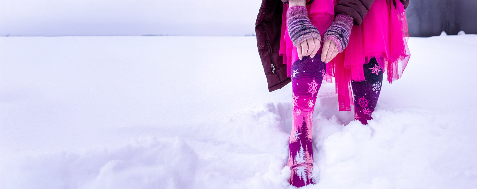 A woman pulls up pink snowflake socks while wearing a pink tulle skirt and standing in the snow.