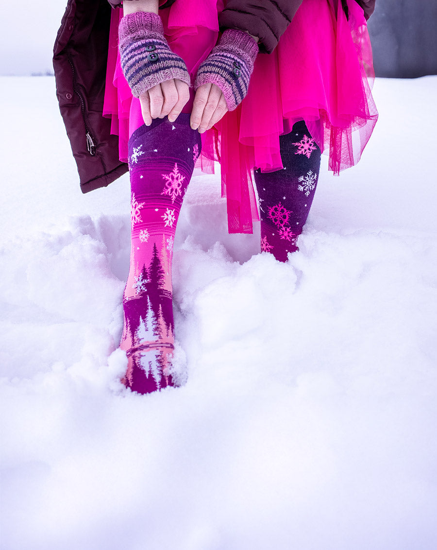 A woman pulls up pink snowflake socks while wearing a pink tulle skirt and standing in the snow.