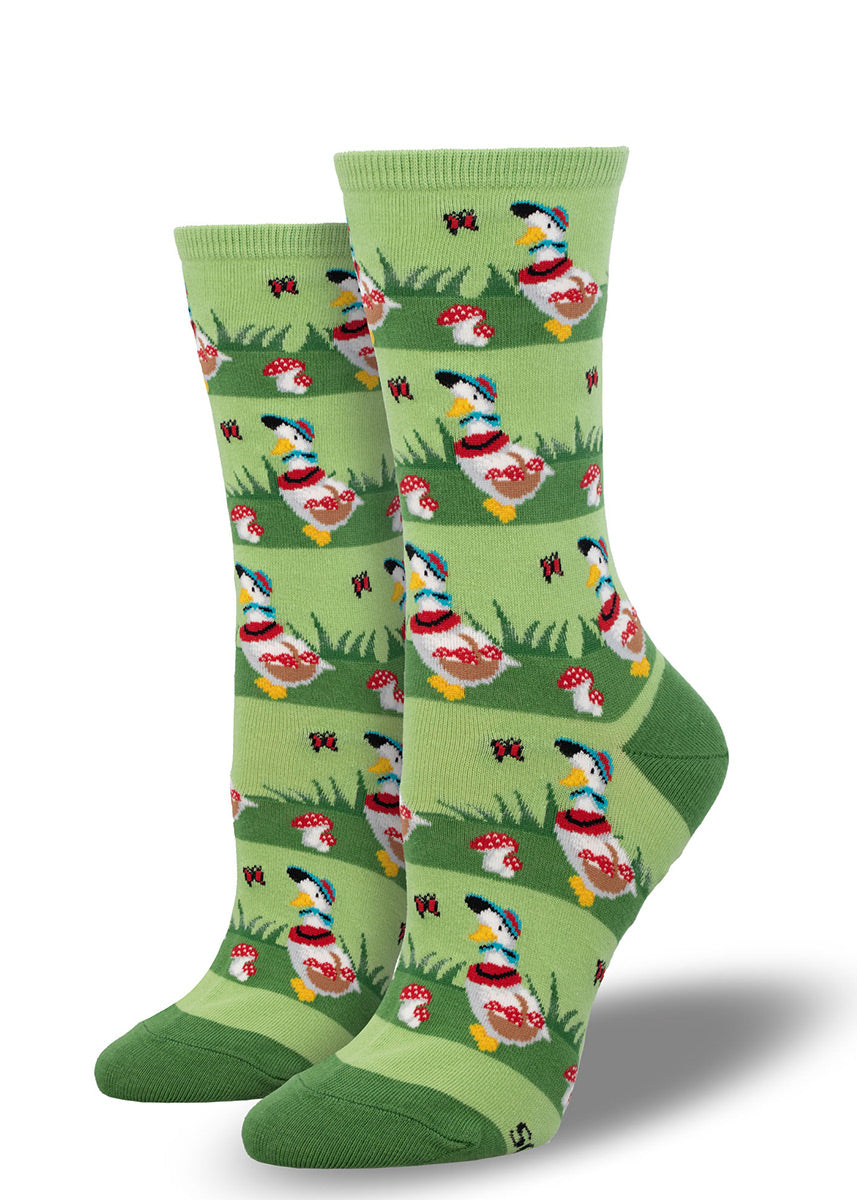 Green crew socks for women with an allover pattern of white geese holding a basket of red mushrooms and standing outside in the grass surrounded by red toadstool mushrooms and butterflies. 