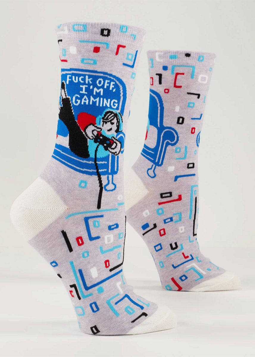 Gray novelty crew socks for women show a cartoon woman laying in a chair holding a video game controller with the words &quot;Fuck Off, I&#39;m Gaming&quot; and an allover geometric pattern in shades of red, blue, black, and white.