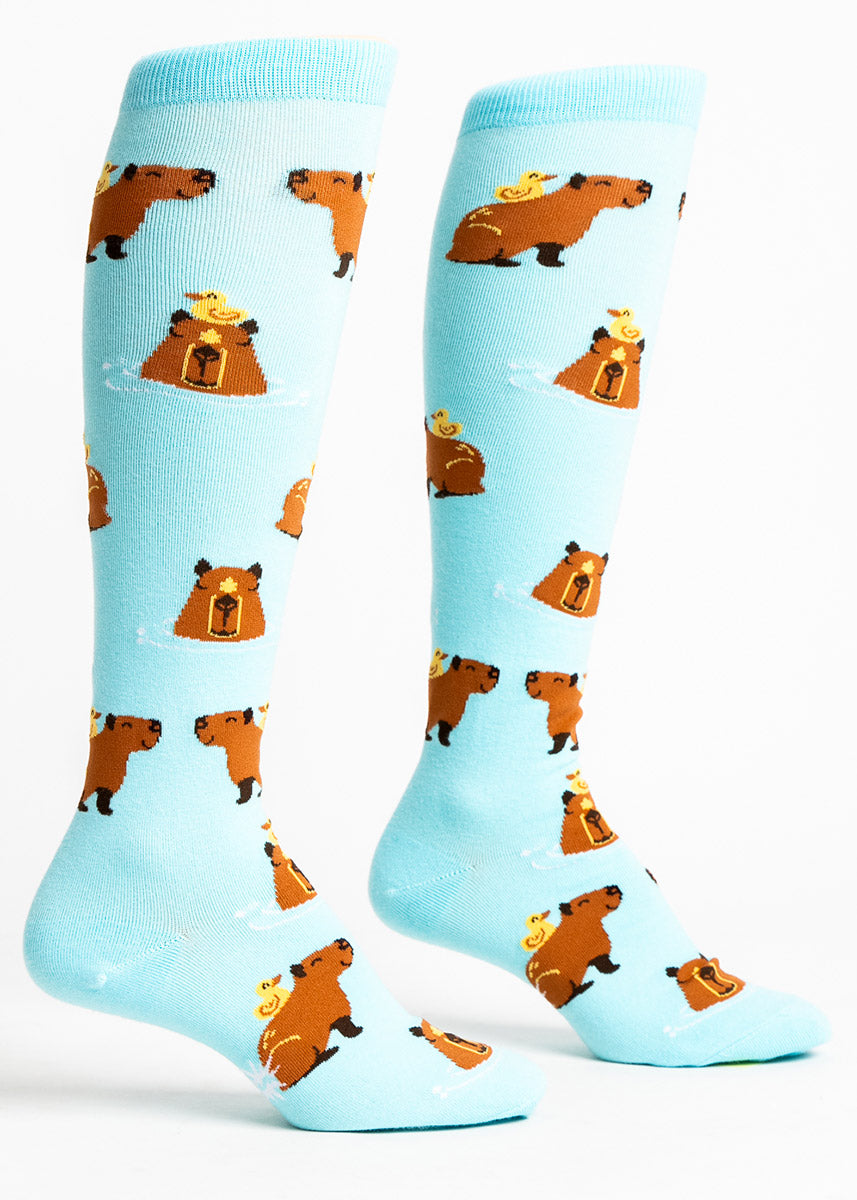 Light blue knee socks for women with a pattern of smiling brown capybara lounging in the water with yellow ducks sitting on their heads and backs.