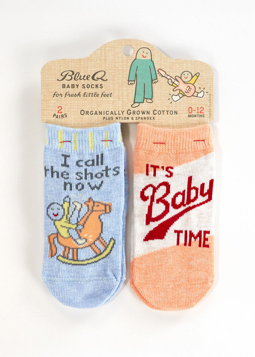 Two pairs of socks for babies: one pair is light blue and features a cartoon baby riding a rocking horse with the words "I call the shots now" and the other has an orange and white color block pattern with the words "It's Baby Time" in red writing.