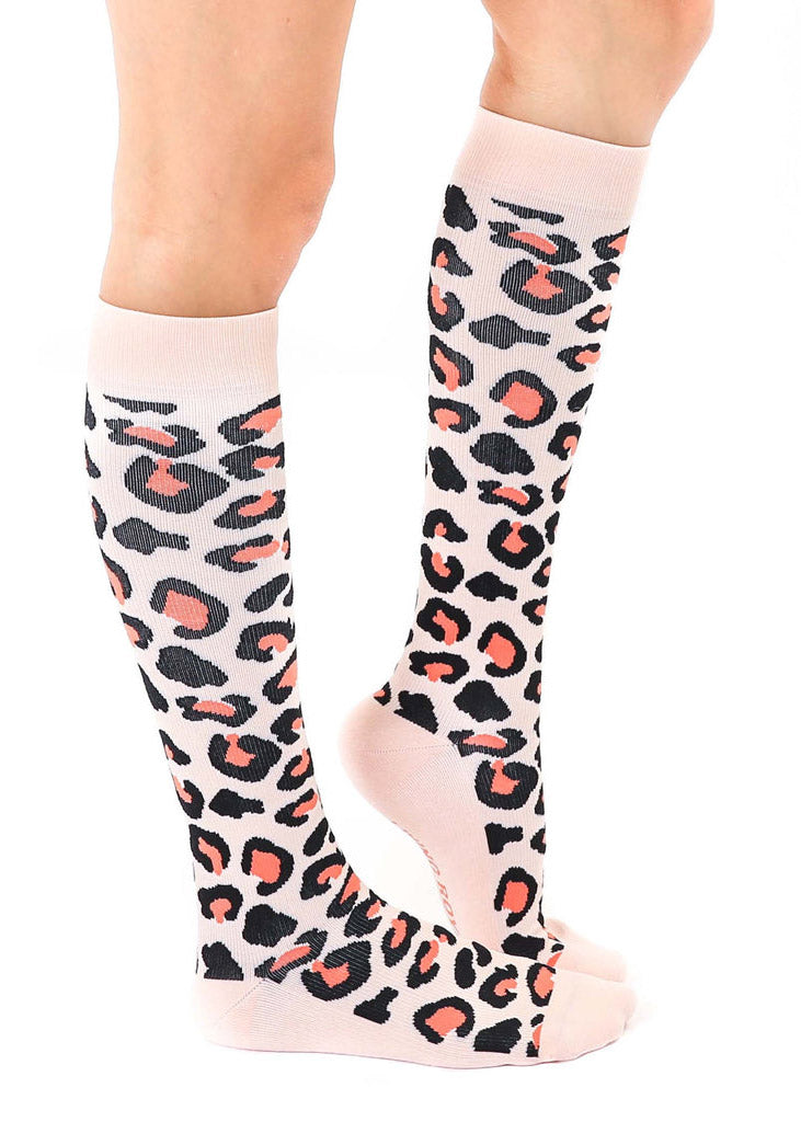 A female model poses straight-on wearing pink knee-high compression socks with an allover orange and black leopard print pattern.