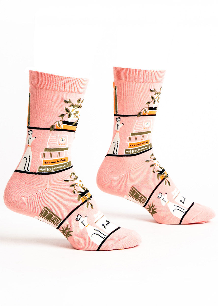 Pink crew socks for women feature horizontal bookshelves with colorful book stacks, plants, and knick-knacks on them.