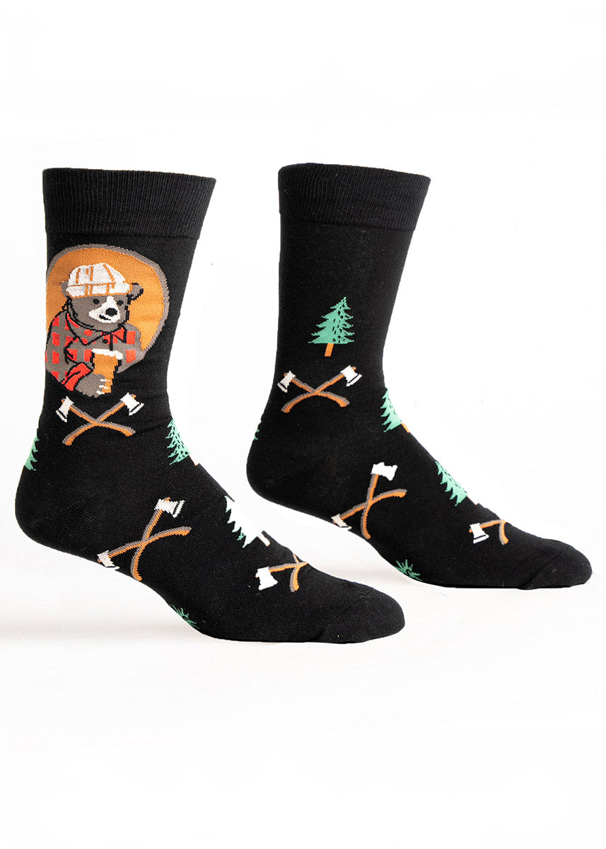 Black novelty crew socks for men with a brown bear dressed as a lumberjack wearing a flannel, beanie, and holding a pint of beer as well as an overall pattern of pine trees and axes. 