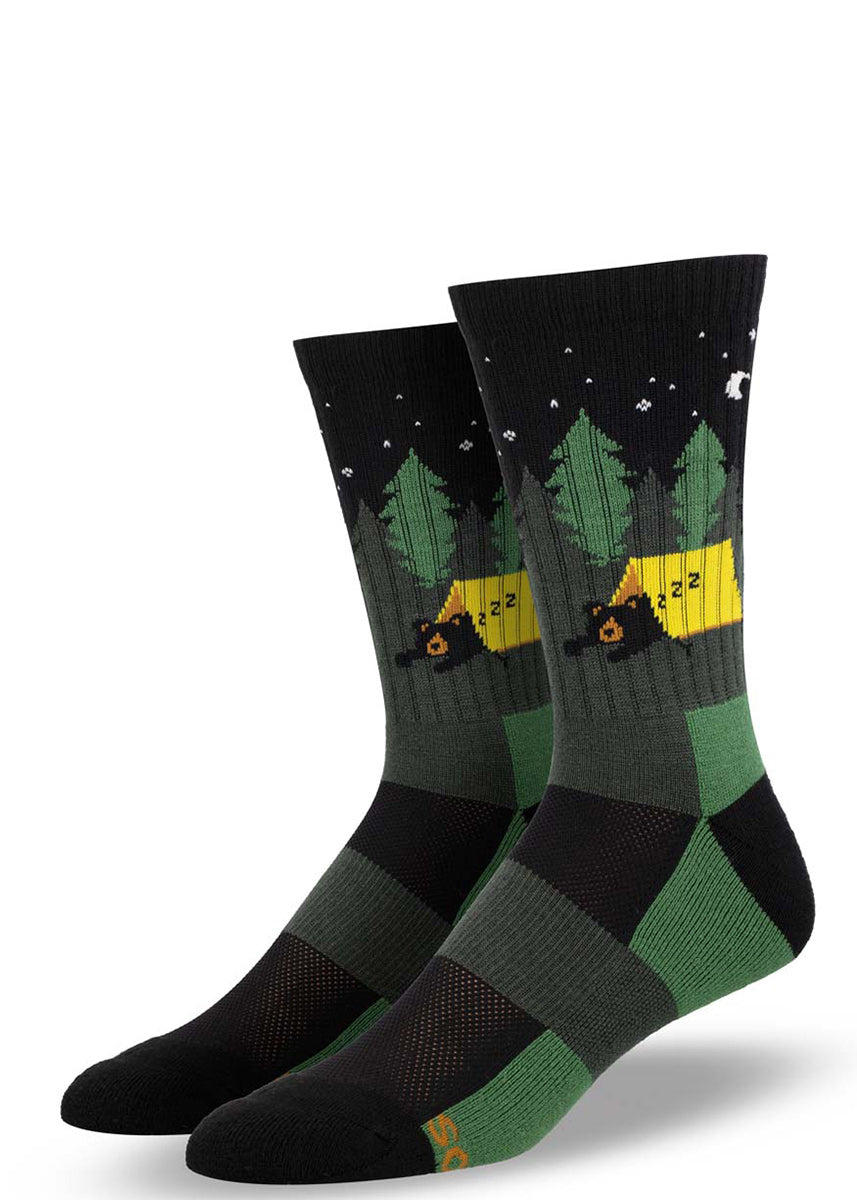 Black and green merino wool hiking socks for men feature a design of a bear sleeping in a yellow tent in starlit woods.