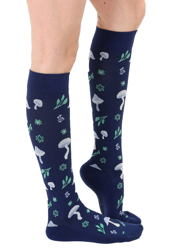 A female model poses straight-on wearing navy knee-high compression socks with an allover pattern of white and light blue toadstool mushrooms and green foliage.