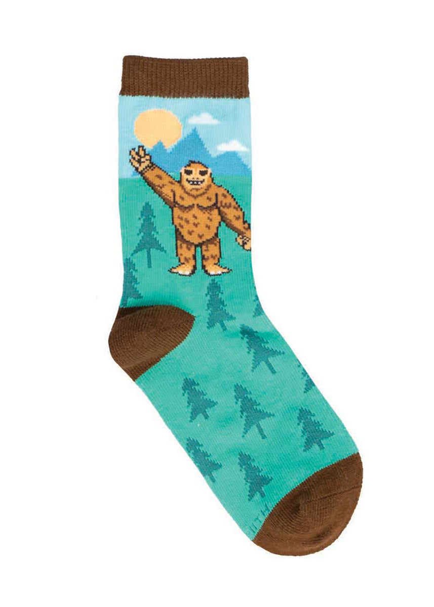 Teal socks for kids that feature a mountain landscape scene along with Bigfoot wearing sunglasses and making a peace sign with his hand. 