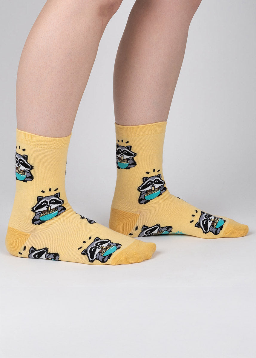 Yellow crew socks for women with an allover pattern of cartoon raccoons eating teal bowls of ramen noodles. 