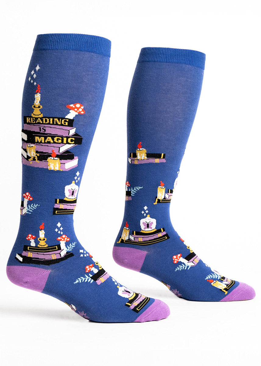 Dark blue knee socks for wide calves that feature a pattern of stacked books, toadstool mushrooms, candles, and stars along with the words &quot;Reading is Magic.&quot; 