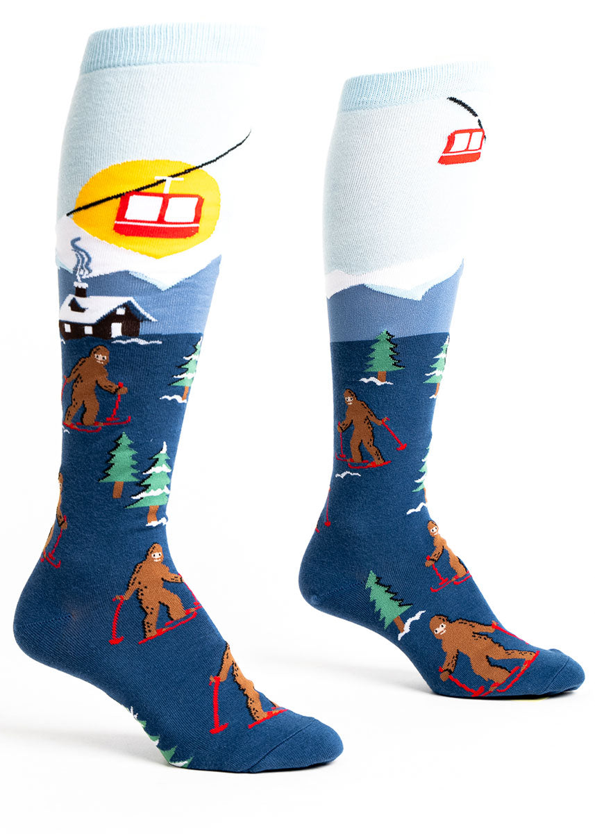 Blue novelty knee socks for women featuring a snowy ski lodge scene with Sasquatches on skis. 