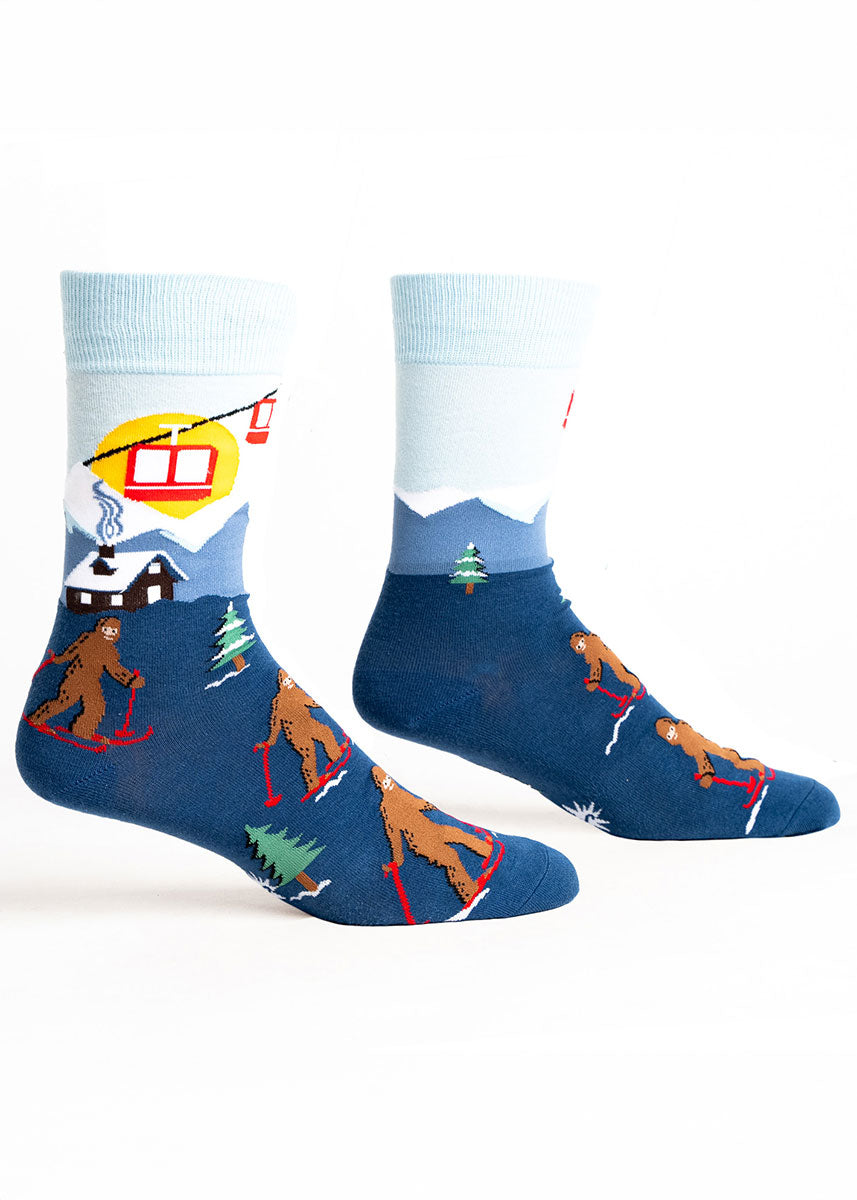 Blue novelty crew socks for men featuring a snowy ski lodge scene with Sasquatches on skis. 