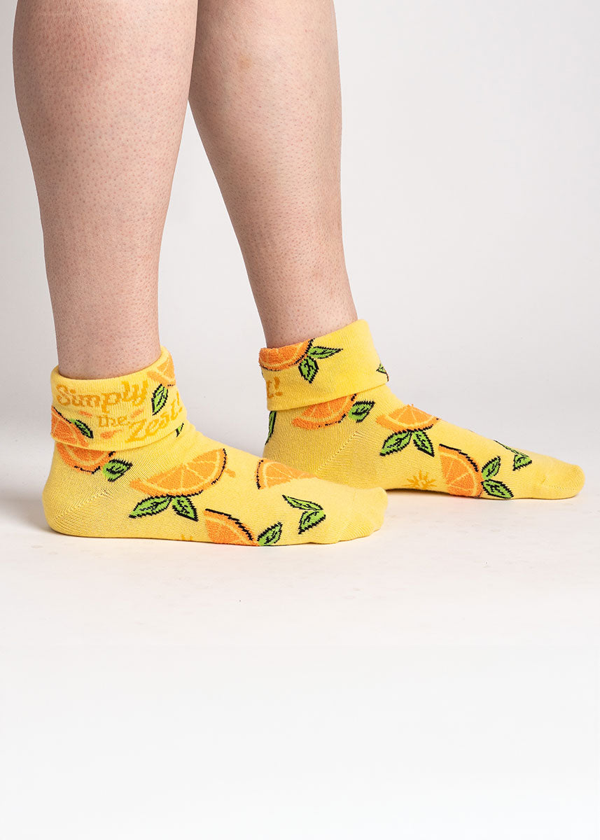 A model poses against a white background wearing yellow turn-cuff ankle socks for women with an allover pattern of fuzzy orange slices and the words &quot;Simply the Zest&quot; written on the cuff.
