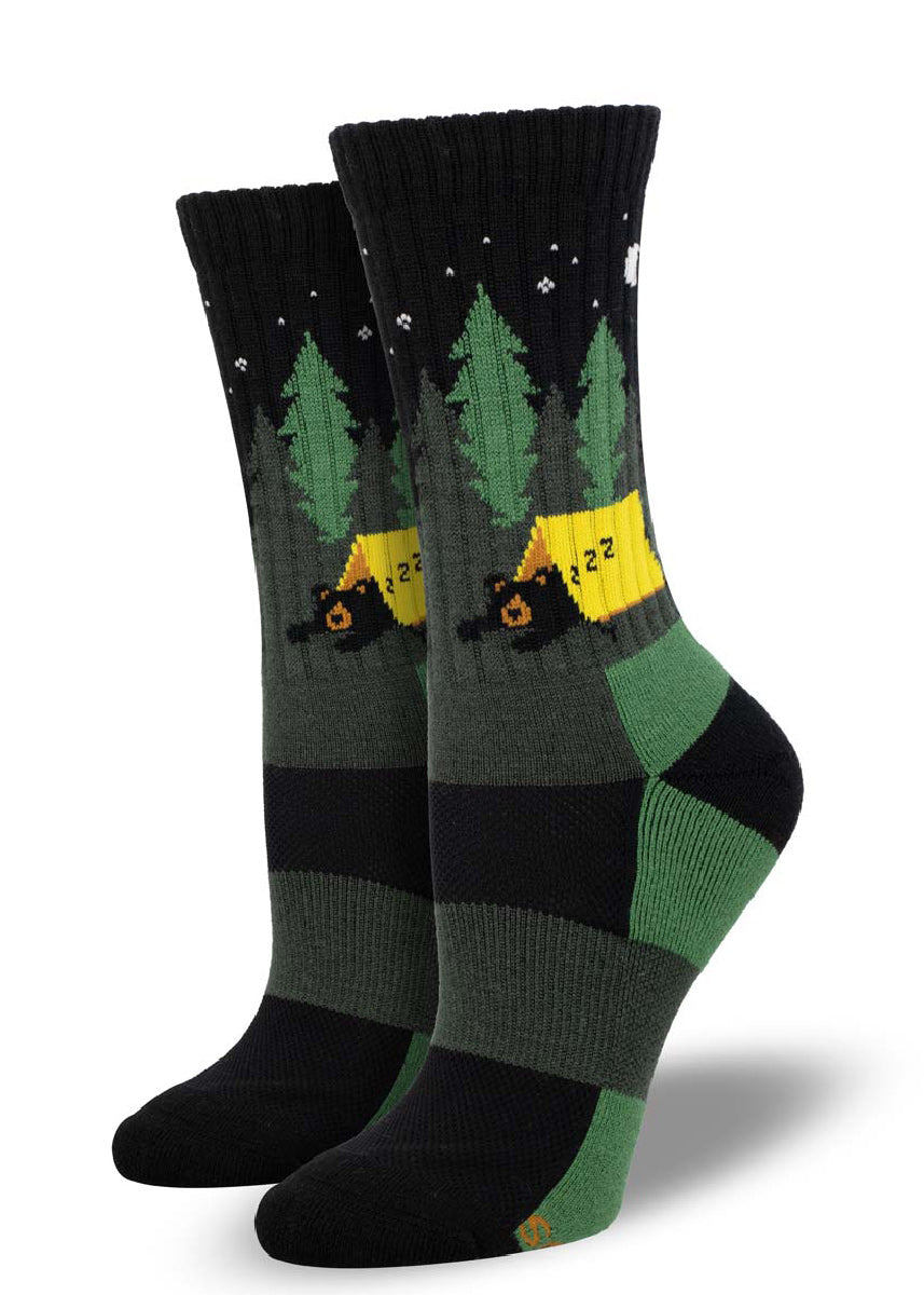 Black and green merino wool hiking socks for women feature a design of a bear sleeping in a yellow tent in starlit woods.